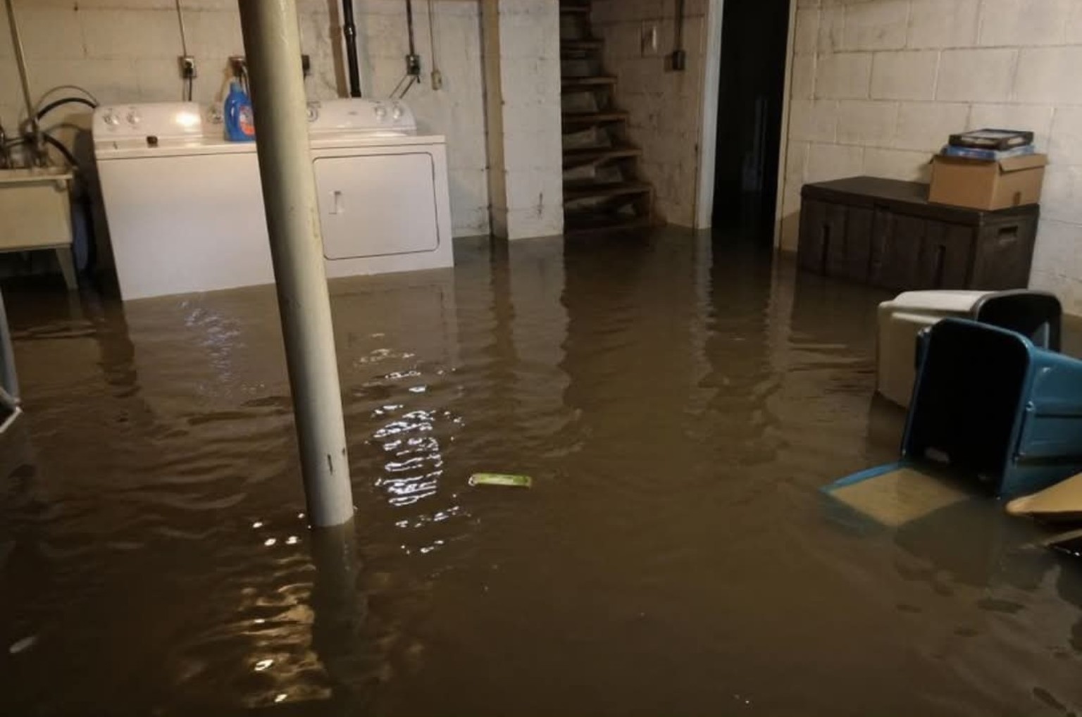 Flooded basement in White House, Tennessee, showing standing water around laundry appliances, stairs, and stored belongings